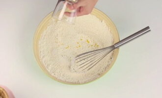 В объемную миску просеиваем муку. Поверх высыпаем указанное количество сахарного песка, соли, дрожжей, ванилина и лимонной цедры. Венчиком смешиваем сухие ингредиенты между собой.